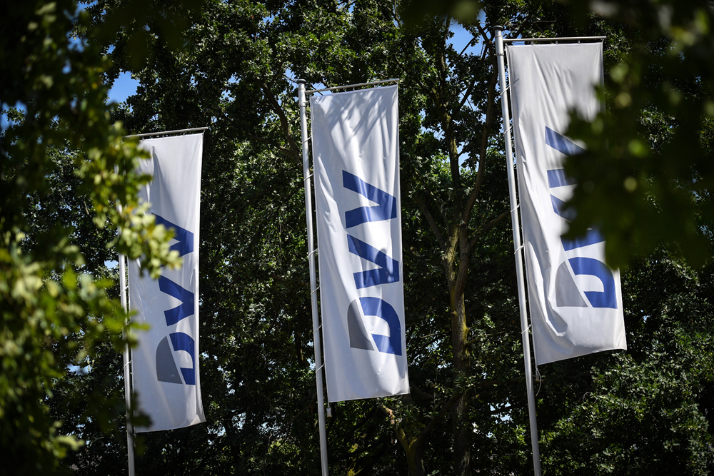 Drei weiße DVV-Fahnen wehen vor grünem Baumbestand im Sonnenlicht.