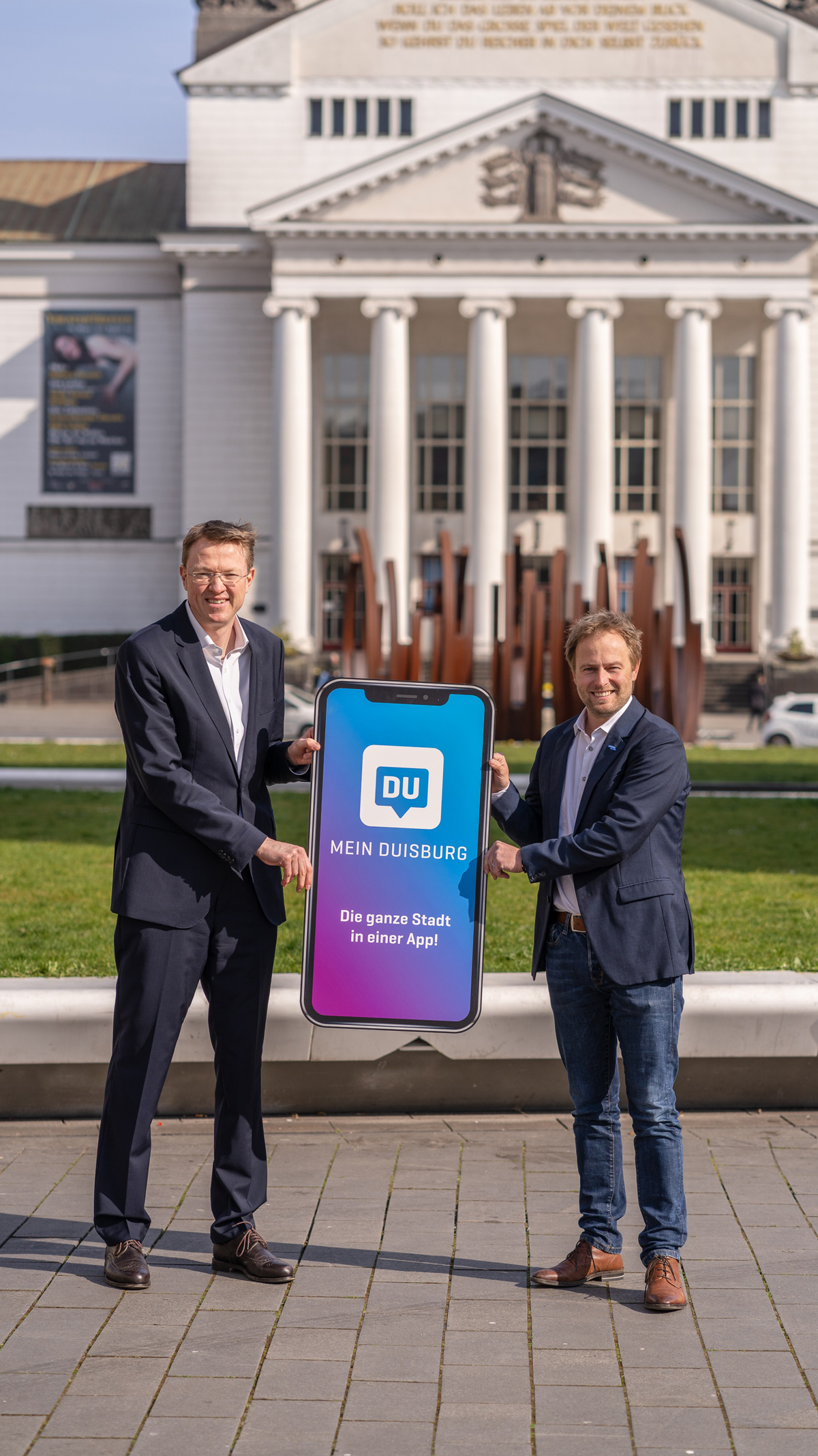 Marcus Vunic und Martin Murrack präsentieren ein übergroßes Smartphone mit der MEIN DUISBURG-App vor dem Stadttheater Duisburg (Hochformat)
