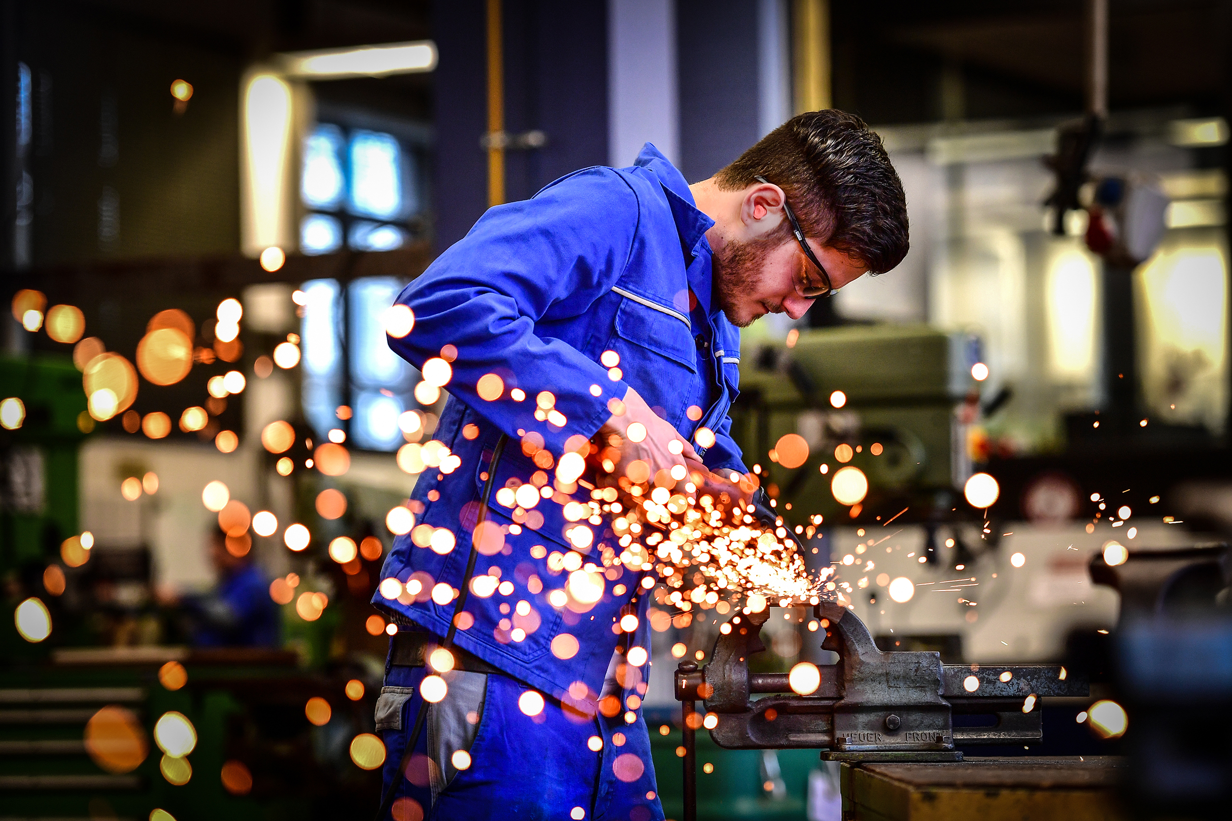 Auszubildender der DVV beim Schleifen eines Metallteils in der Werkstatt, Funken fliegen.